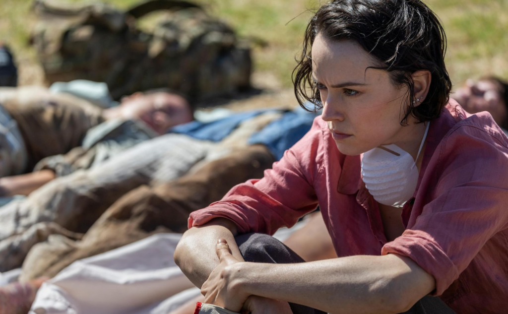 Scene from We Bury the Dead movie review showing a distressed person in a pink shirt with a face mask, sitting outdoors among others lying under blankets, conveying themes of survival, grief, and crisis aftermath.